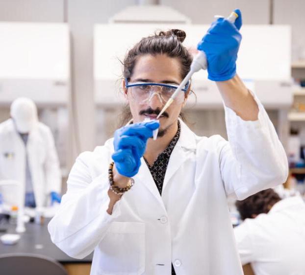 A student wearing a lab coat, protective 眼镜 和 手套 pipettes a sample in a biochemistry lab.