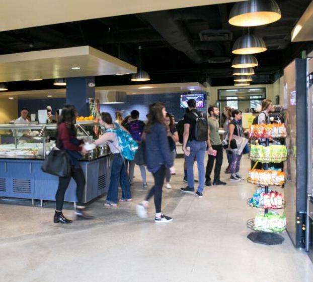 The image shows a busy dining area, likely in a cafeteria. 人们在准备和供应食物的服务站排队. 在左边, there is a refrigerated section stocked with beverages and snacks, and a display with various packaged food items. 气氛热烈。, 和几个学生或赞助人一起, 有些人背着背包, 在空间中移动, 获得食物, 与员工互动.