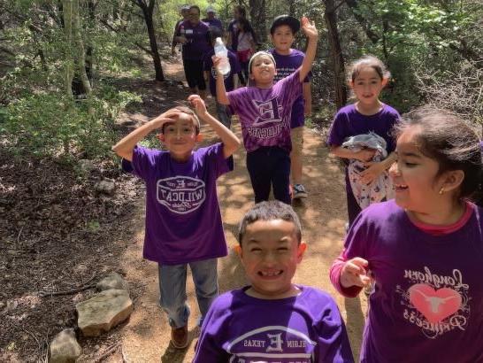 The image shows a group of children on a nature trail. They wear t-shirts with various prints, including "Longhorns Princess" 和 "Wildcats." Trees 和 foliage surround the trail.